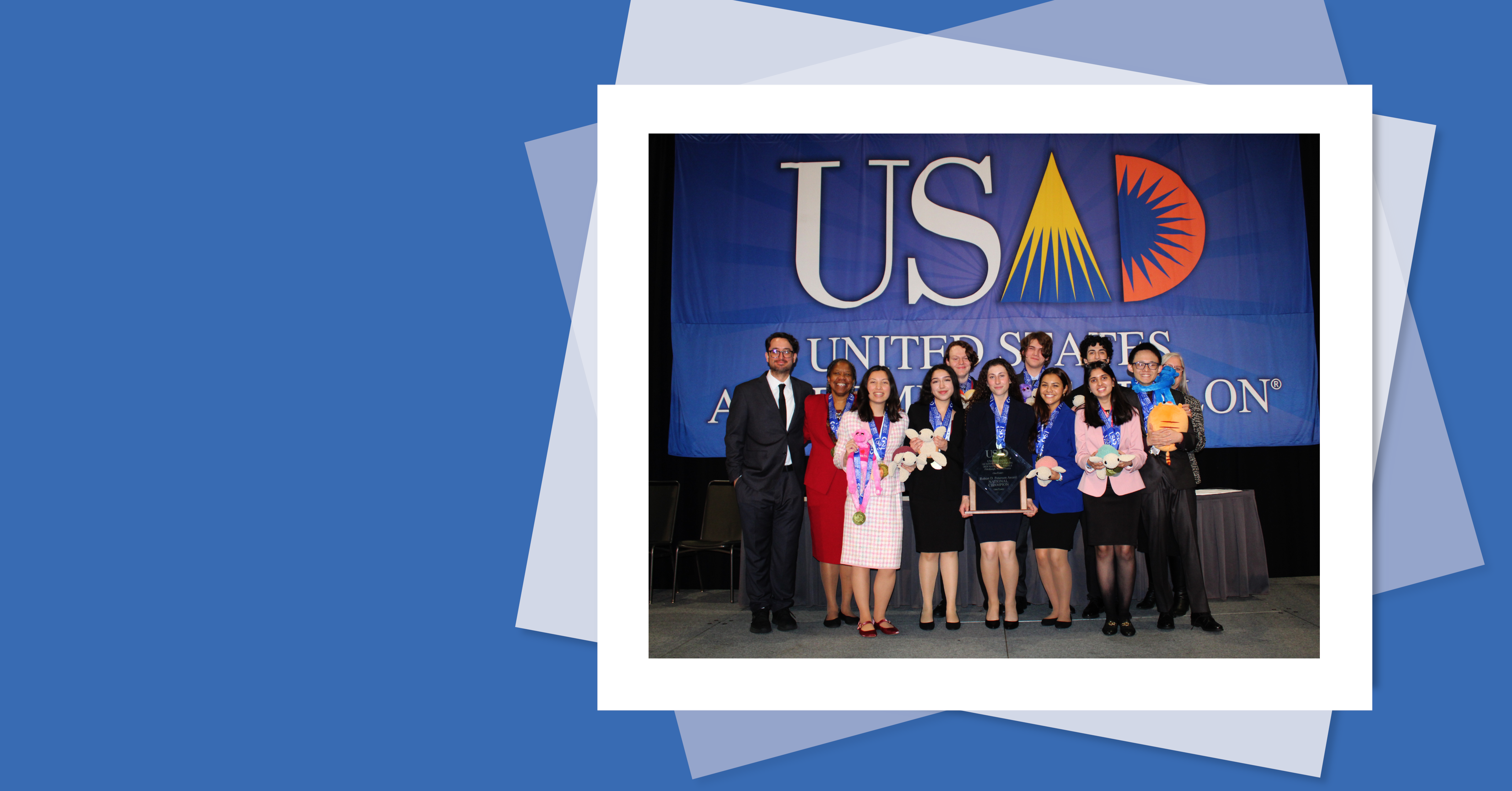  A group of USAD competitors and coaches stand on stage in front of a large United States Academic Decathlon (USAD) banner. They are wearing medals and holding awards, smiling in celebration. The image represents the excitement and achievement of the USAD experience.